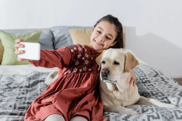 Fröhliches Mädchen macht Selfie auf Handy mit Labrador im Schlafzimmer — Stockfoto