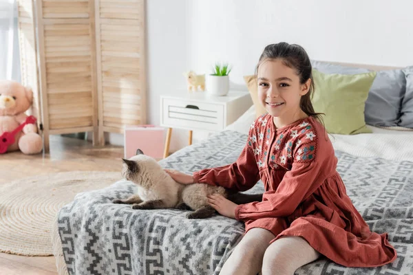 Glückliches Mädchen schaut in die Kamera, während es Katze im Schlafzimmer streichelt — Stockfoto