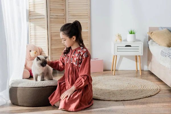 Girl sitting on floor and stroking cat on soft pouf in bedroom — Stock Photo