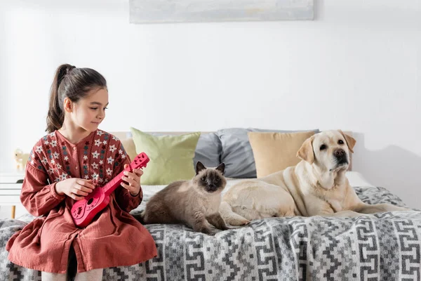 Smiling girl playing toy guitar near cat and labrador dog on bed — Stock Photo