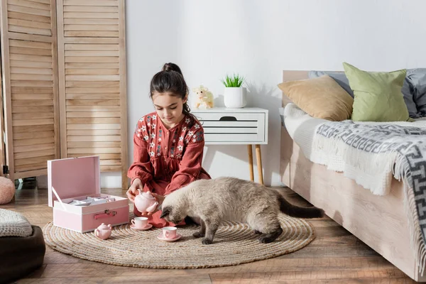 Preteen menina sentada no chão e brincando com brinquedo conjunto de chá e gato — Fotografia de Stock