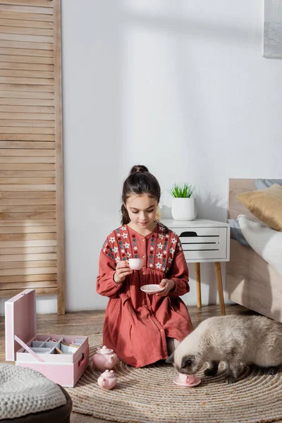 Katze trinkt aus Spielzeugbecher in der Nähe von Mädchen, das auf dem Boden sitzt und mit Spielzeug-Tee-Set spielt — Stockfoto