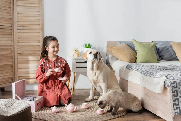 Веселая девушка сидит на полу с игрушечной чашкой, играя рядом с собакой и кошкой — стоковое фото