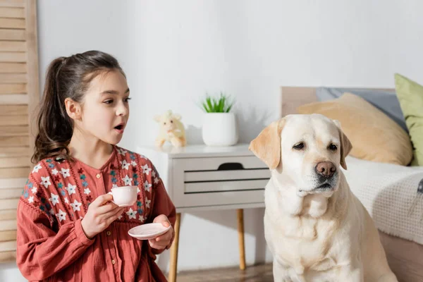Erstauntes Mädchen hält Spielzeugbecher bei gelbem Labrador zu Hause — Stockfoto