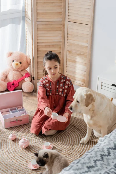 Дівчина показує чашку іграшкового чаю собаці-лабрадору, граючи на підлозі біля набору іграшкового чаю — стокове фото