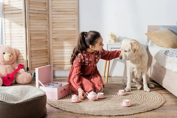 Preteen menina acariciando labrador cão enquanto joga no chão perto de brinquedo conjunto de chá — Fotografia de Stock