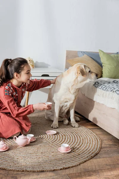 Labrador cão olhando para longe perto menina sentado no chão com copo de chá de brinquedo — Fotografia de Stock