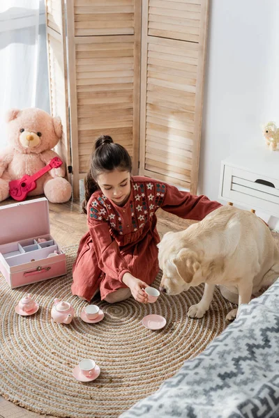 Vue grand angle de fille jouant avec jeu de thé jouet et chien labrador sur le sol à la maison — Photo de stock