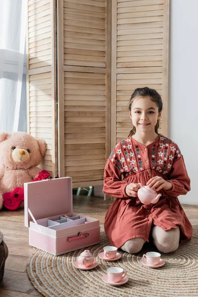 Девочка-подросток сидит на полу рядом с игрушечным чайным сервизом и улыбается перед камерой — стоковое фото