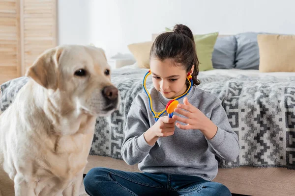 Preteen girl playing doctor with toy stethoscope near blurred labrador dog — Stock Photo