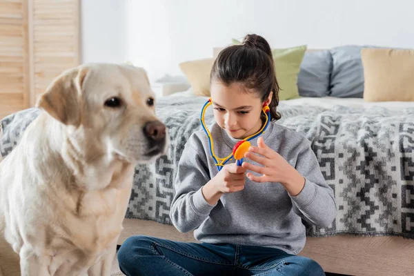 Preteen fille jouer médecin avec jouet stéthoscope près flou labrador chien — Photo de stock