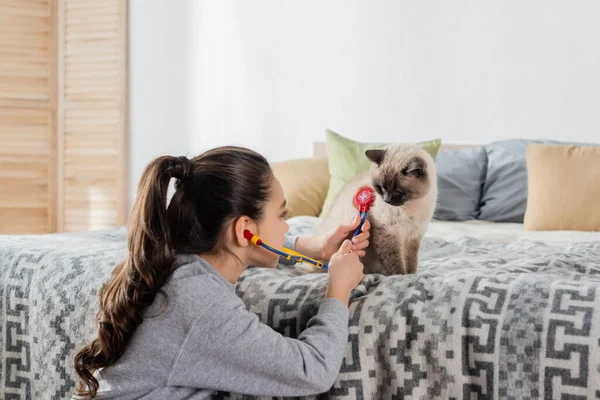 Preteen menina examinando gato com estetoscópio de brinquedo enquanto joga em casa — Fotografia de Stock