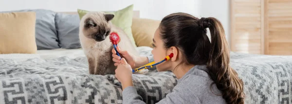 Girl with ponytail examining cat with toy stethoscope at home, banner — Stock Photo