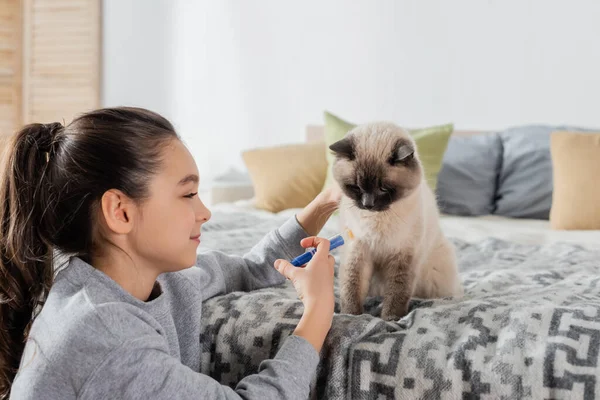 Chica sonriente haciendo inyección a gato con jeringa de juguete en el dormitorio - foto de stock