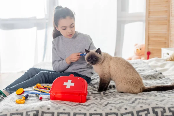 Girl making injection to cat with toy syringe near first aid kit on bed — Stock Photo