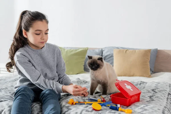 Дівчина сидить на ліжку і грає лікаря з іграшковим медичним набором біля кота — стокове фото