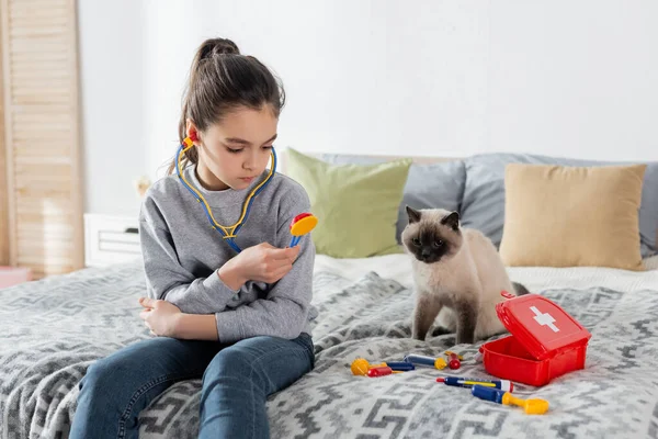 Menina segurando brinquedo estetoscópio enquanto joga na cama perto do gato — Fotografia de Stock