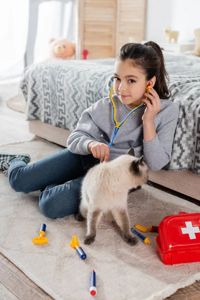 Glückliches Mädchen lächelt in die Kamera, während es Katze mit Spielzeug-Stethoskop auf dem Boden untersucht — Stockfoto