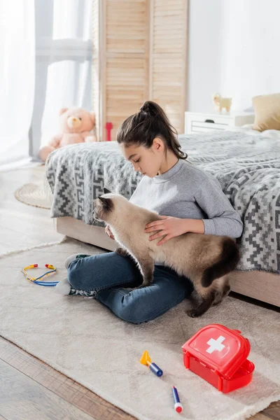 Visão de comprimento total da menina acariciando gato no chão perto de instrumentos médicos de brinquedo — Fotografia de Stock