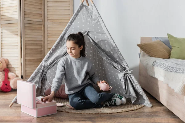Girl holding teapot while sitting in wigwam near box with toy tea set — Stock Photo
