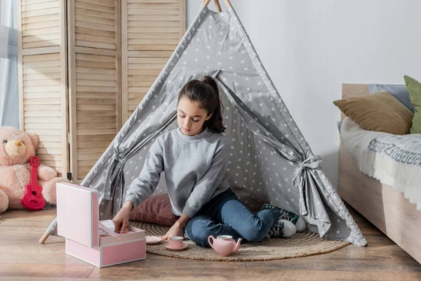 Preteen menina brincando com brinquedo chá definido enquanto sentado no chão em wigwam — Fotografia de Stock