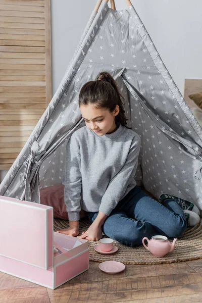 Morena preteen menina sentado no chão em wigwam e jogar com brinquedo chá conjunto — Fotografia de Stock