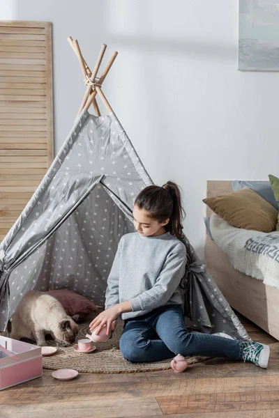 Mädchen gießt Tee aus Spielzeugteekanne, während sie mit Katze in Wigwam spielt — Stockfoto