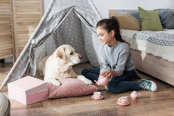 Smiling girl pouring tea from toy teapot near labrador lying in wigwam — Stock Photo