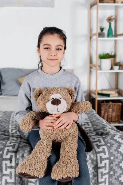 Щаслива дівчина сидить на ліжку з плюшевим ведмедем і дивиться на камеру — стокове фото