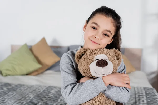 Cheerful preteen girl embracing teddy bear and looking at camera in bedroom — Stock Photo