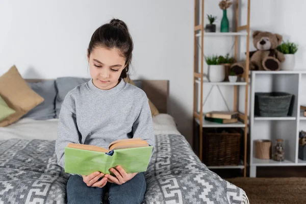 Morena chica preadolescente sentada en la cama y leyendo libro - foto de stock