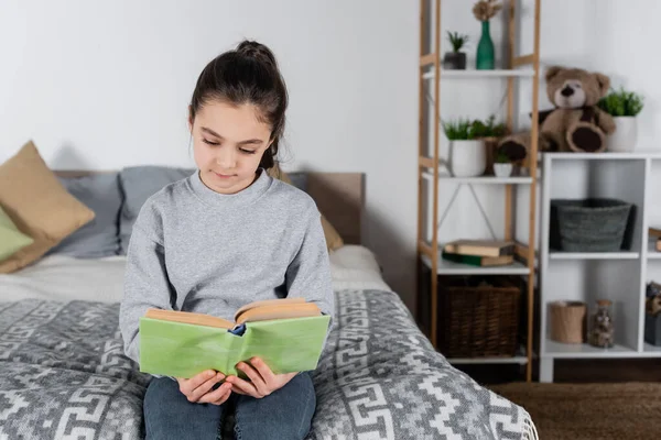 Brünette preteen mädchen sitzen auf bett und lesen buch — Stockfoto