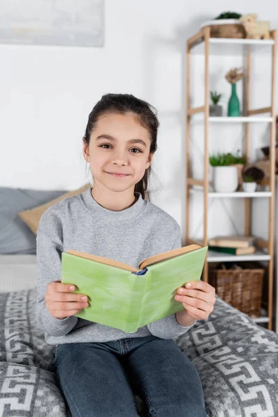 Glückliches brünettes Mädchen lächelt in die Kamera, während es mit Buch im Bett sitzt — Stockfoto