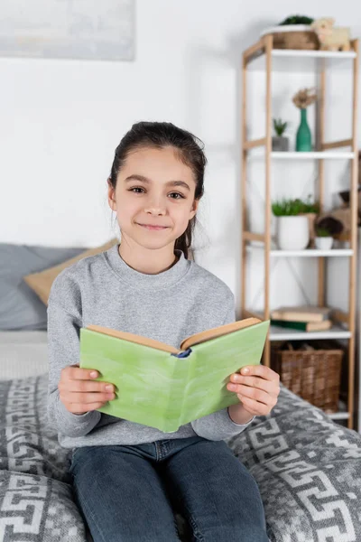 Menina morena feliz sorrindo para a câmera enquanto sentado na cama com livro — Fotografia de Stock