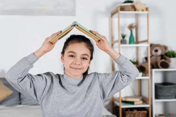 Niño alegre sosteniendo libro sobre la cabeza y sonriendo a la cámara - foto de stock