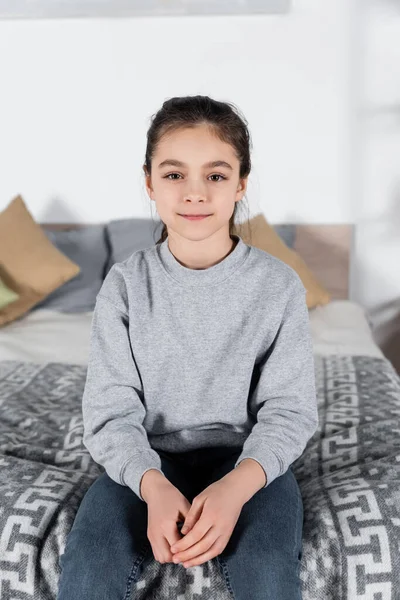 Preteen girl smiling at camera while sitting on blurred bed — Stock Photo