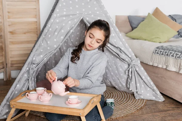 Preteen menina sentado em wigwam e derramando chá de bule de brinquedo — Fotografia de Stock