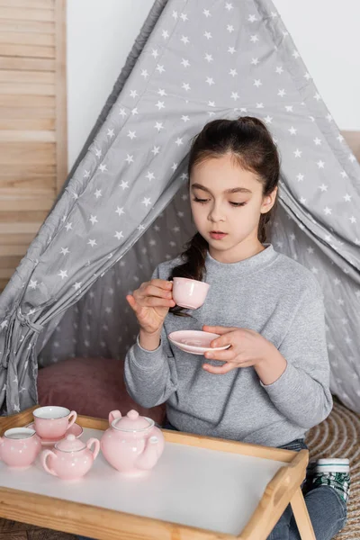 Девушка пьет чай из игрушечной чашки во время игры в вигваме — стоковое фото