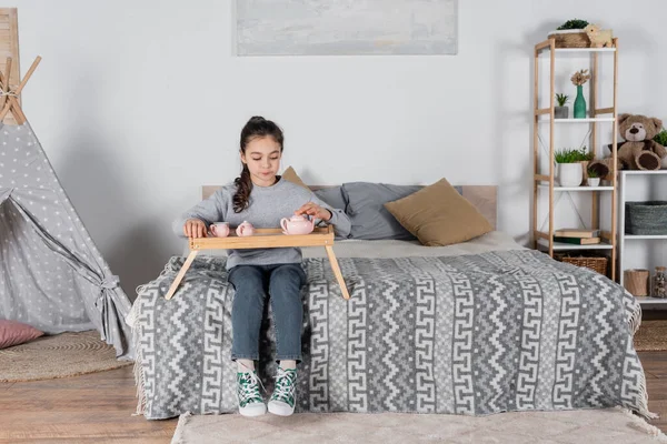 Vista completa della ragazza seduta sul letto con set da tè giocattolo su vassoio di legno — Foto stock