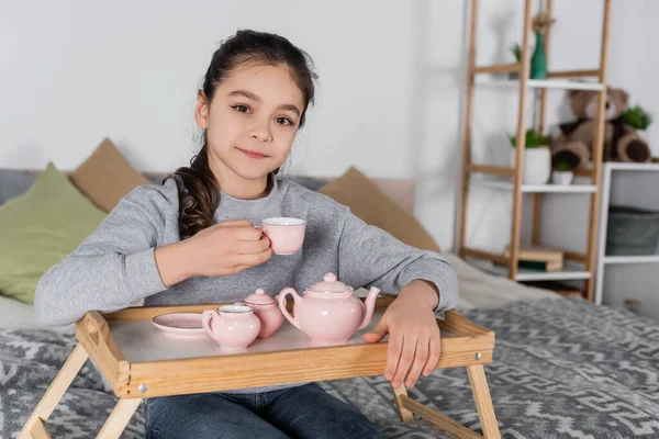 Positives Mädchen blickt in die Kamera, während es Tasse in der Nähe von Spielzeug-Tee-Set hält — Stockfoto