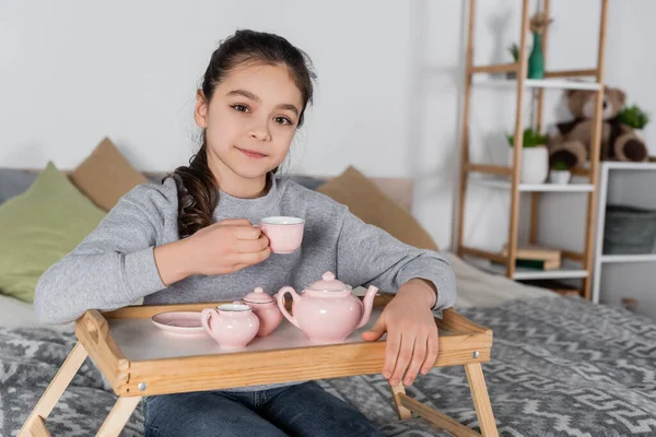 Fille positive regardant la caméra tout en tenant la tasse près du jeu de thé jouet — Photo de stock