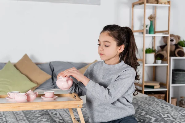 Mädchen gießt Tee aus Spielzeugteekanne, während sie auf dem Bett sitzt — Stockfoto