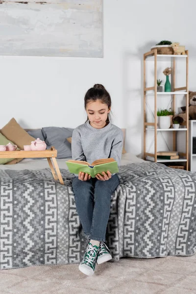 Vista completa de la chica sentada en la cama cerca de la bandeja con juego de té de juguete y libro de lectura - foto de stock