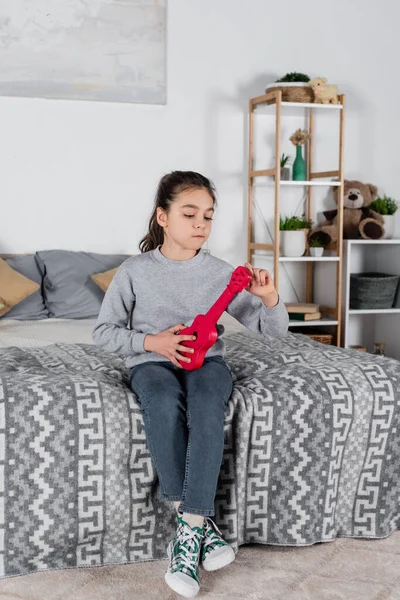 Mädchen stimmt Spielzeuggitarre, während sie zu Hause auf dem Bett sitzt — Stockfoto