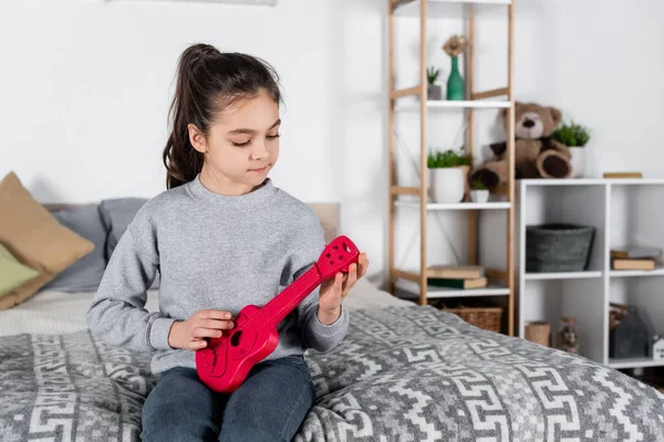Preteen girl sitting in bedroom and playing toy guitar — Stock Photo