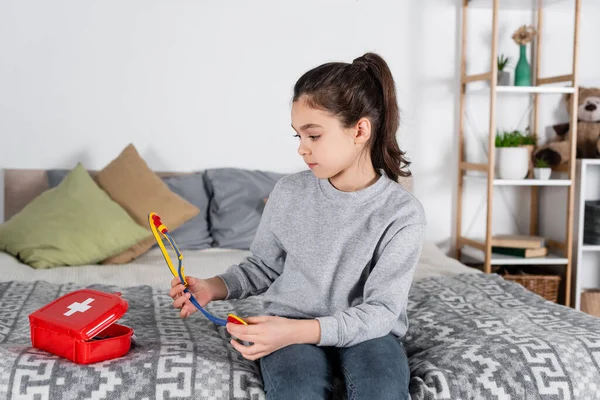 Mädchen sitzt auf Bett neben Verbandskasten und hält Spielzeug-Stethoskop — Stockfoto