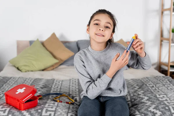Menina sorrindo segurando seringa de brinquedo enquanto sentado na cama perto de kit de primeiros socorros e estetoscópio — Fotografia de Stock
