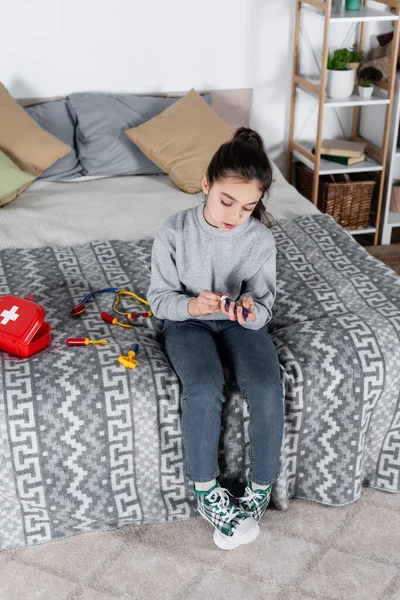 Вид в полный рост девочки с игрушечным шприцем, сидящей на кровати возле медицинского комплекта — стоковое фото