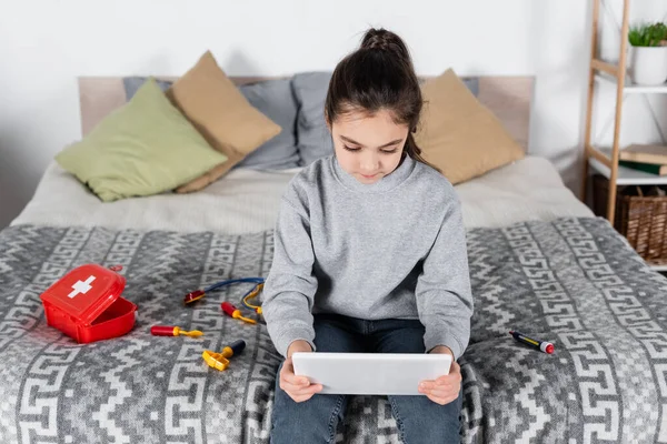 Mädchen benutzt digitales Tablet, während sie auf dem Bett in der Nähe des Spielzeug-medizinischen Sets sitzt — Stockfoto