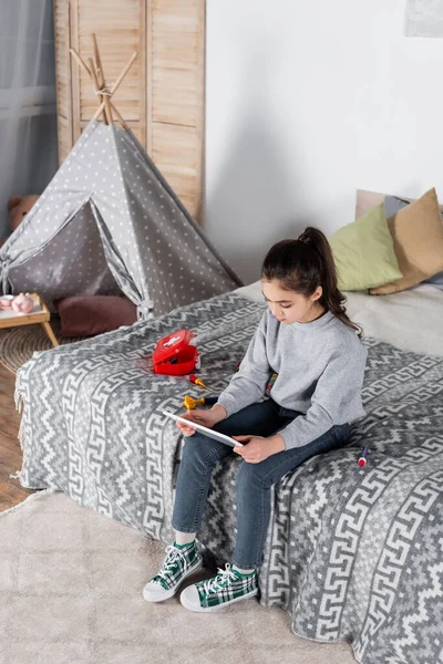 Mädchen mit digitalem Tablet sitzt auf Bett in der Nähe von Spielzeug-Medizinset — Stockfoto